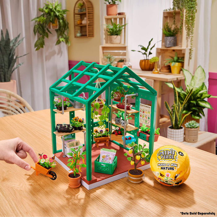 Toy greenhouse set on a table with plants and a hand interacting with it, in a room with plants in the background.